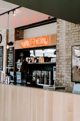 A cozy coffee shop interior with a wooden counter and a neon sign reading 'JUNCTION' mounted above a coffee station. Various coffee-making equipment and bags of coffee beans are neatly arranged on shelves. The walls are made of exposed brick, adding a rustic touch.