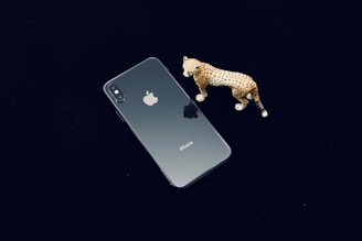 A smartphone with a reflective glass back, featuring the Apple logo, is placed on a dark surface. Next to it, there is a small, detailed figurine of a leopard with a spotted pattern.
