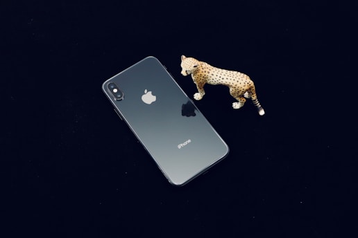 A smartphone with a reflective glass back, featuring the Apple logo, is placed on a dark surface. Next to it, there is a small, detailed figurine of a leopard with a spotted pattern.