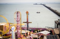 An amusement park by the seaside featuring colorful rides and attractions, including a roller coaster with bright yellow tracks and a tall ride called Sky Drop. A long pier extends over the water, with a boat visible in the sea.