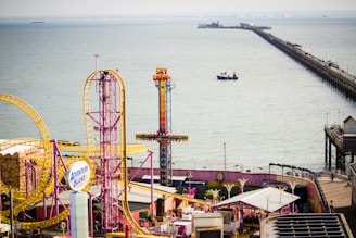An amusement park by the seaside featuring colorful rides and attractions, including a roller coaster with bright yellow tracks and a tall ride called Sky Drop. A long pier extends over the water, with a boat visible in the sea.
