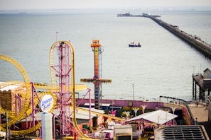 An amusement park by the seaside featuring colorful rides and attractions, including a roller coaster with bright yellow tracks and a tall ride called Sky Drop. A long pier extends over the water, with a boat visible in the sea.