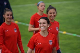 Four women wearing red sports jerseys with a team logo are on a grassy field. They appear to be smiling and possibly engaged in a sports activity or training session.