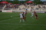 A women's soccer match is taking place, with players from two teams wearing different uniforms, one predominantly in white and the other in red and black. The action is centered on a player in white who appears to be attempting to head the ball, while a player in red and black is closely competing. The stadium is filled with spectators in the background, and there are many banners and advertisements visible around the field.
