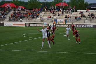 A women's soccer match is taking place, with players from two teams wearing different uniforms, one predominantly in white and the other in red and black. The action is centered on a player in white who appears to be attempting to head the ball, while a player in red and black is closely competing. The stadium is filled with spectators in the background, and there are many banners and advertisements visible around the field.