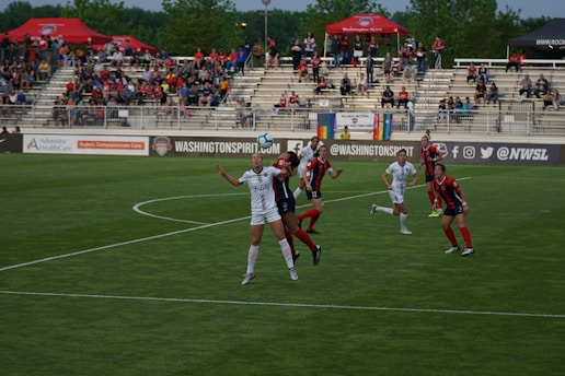 A women's soccer match is taking place, with players from two teams wearing different uniforms, one predominantly in white and the other in red and black. The action is centered on a player in white who appears to be attempting to head the ball, while a player in red and black is closely competing. The stadium is filled with spectators in the background, and there are many banners and advertisements visible around the field.