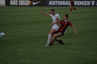 Two soccer players are engaged in a dynamic action on a grassy field. One player, wearing a white uniform, is in a defensive stance while the other, in a red and black uniform, appears to be dribbling or shielding the ball. The background shows a blurred advertisement for Nike Soccer and signage for a women's league.