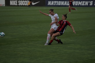 Two soccer players are engaged in a dynamic action on a grassy field. One player, wearing a white uniform, is in a defensive stance while the other, in a red and black uniform, appears to be dribbling or shielding the ball. The background shows a blurred advertisement for Nike Soccer and signage for a women's league.