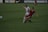 Two soccer players are engaged in a dynamic action on a grassy field. One player, wearing a white uniform, is in a defensive stance while the other, in a red and black uniform, appears to be dribbling or shielding the ball. The background shows a blurred advertisement for Nike Soccer and signage for a women's league.