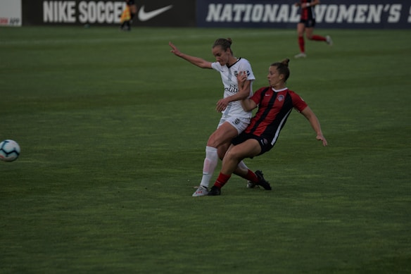 Two soccer players are engaged in a dynamic action on a grassy field. One player, wearing a white uniform, is in a defensive stance while the other, in a red and black uniform, appears to be dribbling or shielding the ball. The background shows a blurred advertisement for Nike Soccer and signage for a women's league.