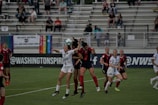 A group of female soccer players jump to head the ball during a match. The players are wearing uniforms, with one team in black and red stripes and the other in white. In the background, there are spectators seated in the stands, along with various flags displayed, including a rainbow flag.