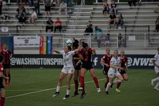 A group of female soccer players jump to head the ball during a match. The players are wearing uniforms, with one team in black and red stripes and the other in white. In the background, there are spectators seated in the stands, along with various flags displayed, including a rainbow flag.