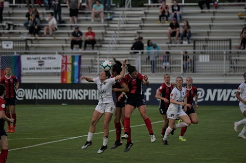 A group of female soccer players jump to head the ball during a match. The players are wearing uniforms, with one team in black and red stripes and the other in white. In the background, there are spectators seated in the stands, along with various flags displayed, including a rainbow flag.