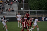 A group of female soccer players from two opposing teams are actively playing on a well-maintained green field. The players are wearing white and red kits, and they are jumping and reaching to head or kick a soccer ball in mid-air. The scene is dynamic and intense, capturing the action of a competitive match. Spectators can be seen seated in the background, watching the game with interest.