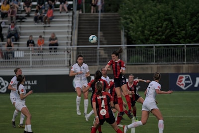 A group of female soccer players from two opposing teams are actively playing on a well-maintained green field. The players are wearing white and red kits, and they are jumping and reaching to head or kick a soccer ball in mid-air. The scene is dynamic and intense, capturing the action of a competitive match. Spectators can be seen seated in the background, watching the game with interest.