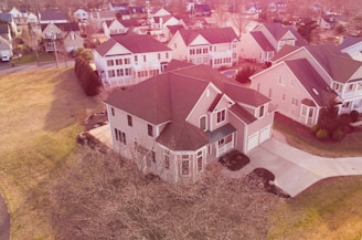 A suburban neighborhood with a bird's-eye view showing multiple houses. The central focus is on a large two-story house with a brown roof and cream-colored exterior. The house is surrounded by a well-maintained lawn and a paved driveway. Other similar houses are visible in the background, along with trees that have shed their leaves, indicating a fall or early winter season.