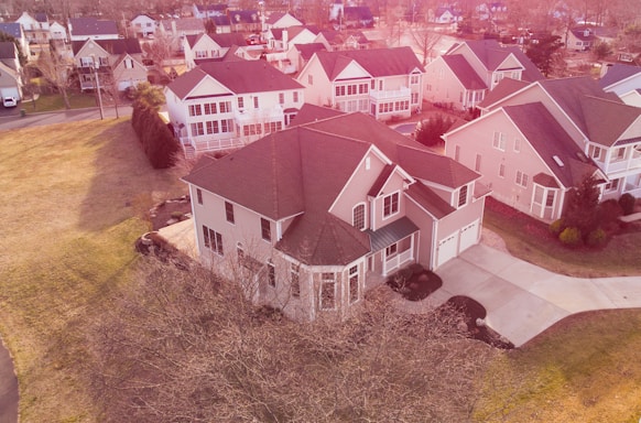 A suburban neighborhood with a bird's-eye view showing multiple houses. The central focus is on a large two-story house with a brown roof and cream-colored exterior. The house is surrounded by a well-maintained lawn and a paved driveway. Other similar houses are visible in the background, along with trees that have shed their leaves, indicating a fall or early winter season.