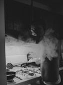 A person is cooking on a gas stove, surrounded by a cloud of steam. The scene is in black and white, giving it a moody and dramatic feel. The person appears to be concentrating on what they are doing, with their hand reaching towards a pan using a slotted spoon.