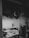 A person is cooking on a gas stove, surrounded by a cloud of steam. The scene is in black and white, giving it a moody and dramatic feel. The person appears to be concentrating on what they are doing, with their hand reaching towards a pan using a slotted spoon.