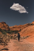 A lone hiker with trekking poles walks along a rugged, rocky path through a canyon with vibrant red rock formations under a clear blue sky. A solitary cloud floats overhead, adding contrast to the scene.
