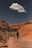 A lone hiker with trekking poles walks along a rugged, rocky path through a canyon with vibrant red rock formations under a clear blue sky. A solitary cloud floats overhead, adding contrast to the scene.