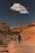 A lone hiker with trekking poles walks along a rugged, rocky path through a canyon with vibrant red rock formations under a clear blue sky. A solitary cloud floats overhead, adding contrast to the scene.
