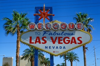 A vibrant sign with the words 'Welcome to Fabulous Las Vegas Nevada' is prominently displayed. The sign features a retro design with illuminated letters and a starburst at the top. It is flanked by tall palm trees, set against a clear blue sky with some power lines visible in the background.