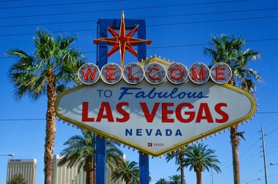 A vibrant sign with the words 'Welcome to Fabulous Las Vegas Nevada' is prominently displayed. The sign features a retro design with illuminated letters and a starburst at the top. It is flanked by tall palm trees, set against a clear blue sky with some power lines visible in the background.
