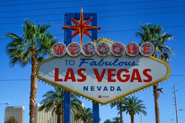 A vibrant sign with the words 'Welcome to Fabulous Las Vegas Nevada' is prominently displayed. The sign features a retro design with illuminated letters and a starburst at the top. It is flanked by tall palm trees, set against a clear blue sky with some power lines visible in the background.