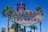 A vibrant sign with the words 'Welcome to Fabulous Las Vegas Nevada' is prominently displayed. The sign features a retro design with illuminated letters and a starburst at the top. It is flanked by tall palm trees, set against a clear blue sky with some power lines visible in the background.