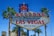 A vibrant sign with the words 'Welcome to Fabulous Las Vegas Nevada' is prominently displayed. The sign features a retro design with illuminated letters and a starburst at the top. It is flanked by tall palm trees, set against a clear blue sky with some power lines visible in the background.