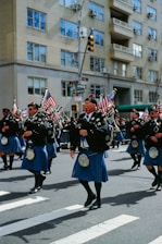 A group of people dressed in traditional Scottish attire, including kilts and sporrans, are marching down a city street. They are playing bagpipes and carrying American flags. The background shows a cityscape with an apartment building and traffic signals.