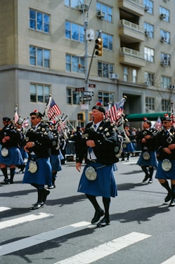 A group of people dressed in traditional Scottish attire, including kilts and sporrans, are marching down a city street. They are playing bagpipes and carrying American flags. The background shows a cityscape with an apartment building and traffic signals.