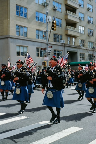 A group of people dressed in traditional Scottish attire, including kilts and sporrans, are marching down a city street. They are playing bagpipes and carrying American flags. The background shows a cityscape with an apartment building and traffic signals.
