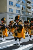 Brightly uniformed bagpipe players march in formation across a crosswalk on a sunny day. They are wearing traditional Scottish attire with yellow kilts and navy blue jackets, topped with tall black hats and yellow plumes. The background features a city street with residential buildings, reflecting an urban parade setting.