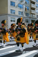 Brightly uniformed bagpipe players march in formation across a crosswalk on a sunny day. They are wearing traditional Scottish attire with yellow kilts and navy blue jackets, topped with tall black hats and yellow plumes. The background features a city street with residential buildings, reflecting an urban parade setting.