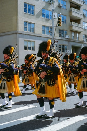 Brightly uniformed bagpipe players march in formation across a crosswalk on a sunny day. They are wearing traditional Scottish attire with yellow kilts and navy blue jackets, topped with tall black hats and yellow plumes. The background features a city street with residential buildings, reflecting an urban parade setting.