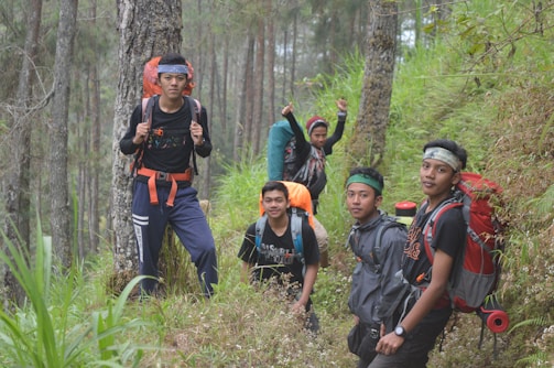 Five young men are hiking through a dense forest, wearing outdoor gear including backpacks and headbands. The scene is lush with greenery, and the group appears to be on a narrow trail surrounded by tall trees and plants. The hikers are dressed in sporty attire, indicating a journey in nature.
