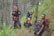Five young men are hiking through a dense forest, wearing outdoor gear including backpacks and headbands. The scene is lush with greenery, and the group appears to be on a narrow trail surrounded by tall trees and plants. The hikers are dressed in sporty attire, indicating a journey in nature.