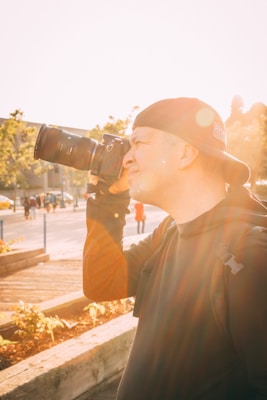 A person wearing a backward cap is using a DSLR camera to take a photo in an outdoor setting with the sun shining brightly, causing lens flare. The background features trees, foliage, and people walking at a distance.