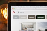 Close-up view of a laptop screen displaying the homepage of a Pinterest website, with various category buttons such as Room, Houseplant, Inspiration, and Garden Ideas. The browser interface is visible with tabs and a search bar, and part of a lit light bulb is seen in the blurred background, adding a warm glow.