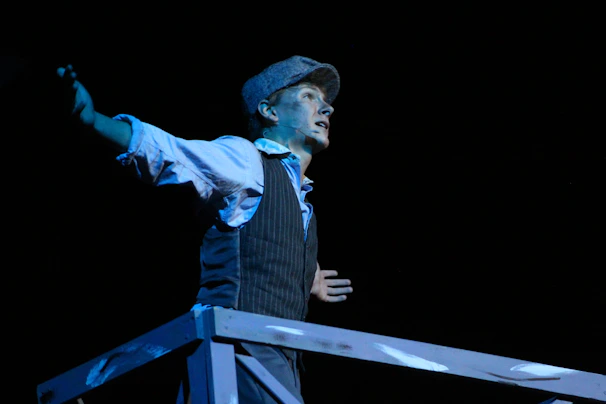A person dressed in an old-fashioned outfit, wearing a vest and a cap, passionately performing on stage. Their arms are outstretched, and they appear to be singing or speaking dramatically.