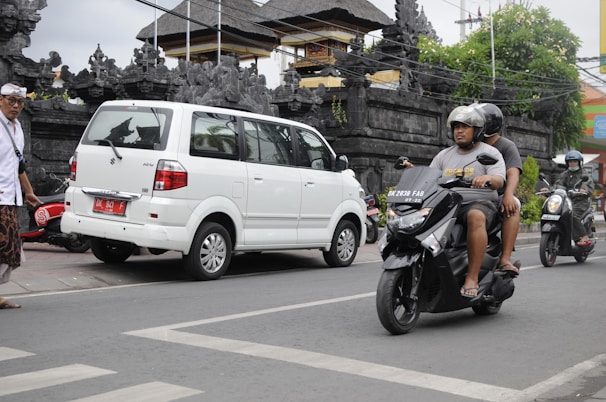 In a street setting, two individuals on a black scooter are seen riding past a parked white van. The background features traditional stone structures and greenery, suggesting a blend of modern and cultural elements. A few other scooters and a person wearing traditional attire are visible.
