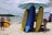 A beach scene featuring several colorful surfboards propped up under large umbrellas on sandy ground. People are relaxing and enjoying the sun near the shoreline, with the ocean visible in the background.