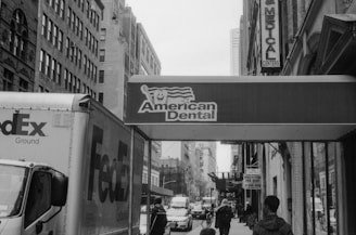 A bustling urban street scene features a FedEx truck parked alongside the sidewalk. Buildings lined with large windows frame the narrow street, which is populated with pedestrians. A prominent sign for 'American Dental' hangs above, suggesting the presence of a dental office nearby. Traffic and city life are visibly active in the background.