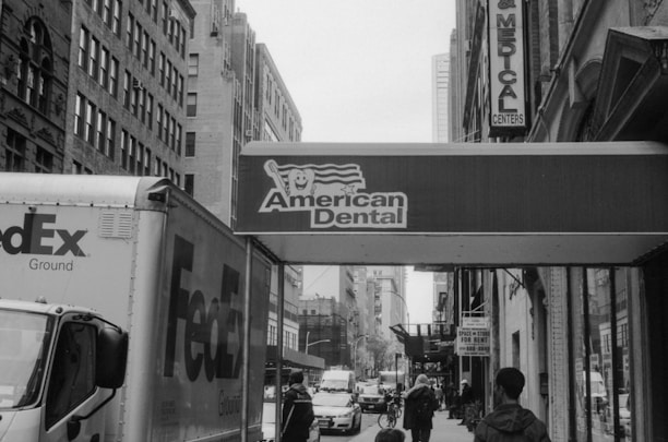 A bustling urban street scene features a FedEx truck parked alongside the sidewalk. Buildings lined with large windows frame the narrow street, which is populated with pedestrians. A prominent sign for 'American Dental' hangs above, suggesting the presence of a dental office nearby. Traffic and city life are visibly active in the background.