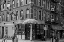 A street corner in an urban setting features a brick building with a striped awning marked 'Williamsburg Pizza' on the ground floor. Above the storefront, the building has multiple windows with decorative trim. A street sign for Broome Street is visible, along with traffic lights and people walking nearby. Bicycles are parked against the building, and fire escapes are attached to the upper floors.