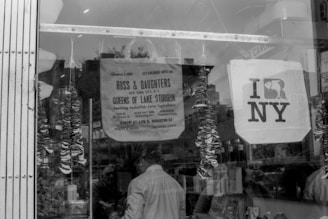 A black and white photograph of a storefront window displaying various items. There is a sign for Russ & Daughters, which is known for smoked fish and other deli goods. Another sign visible is an 'I love NY' poster. The window is adorned with intricate decorations and reflections of people can be seen.