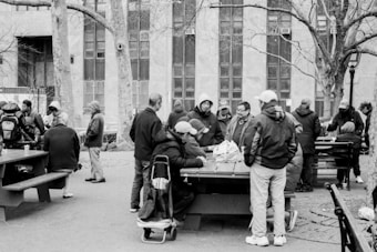A group of people are gathered around large picnic tables in an outdoor urban area. They are engaged in conversation and activities such as playing games. The trees in the area are bare, suggesting it's a cold season. A large building with multiple windows forms the backdrop. Most individuals are wearing jackets and hoodies, indicating chilly weather.