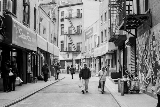 A black and white photograph of a narrow urban street with several people walking or standing. There are various small shops with awnings lining both sides of the street. Fire escapes are visible on the buildings, and graffiti or murals are painted on one of the walls. Some individuals are carrying bags, and the setting appears to be a typical city neighborhood.